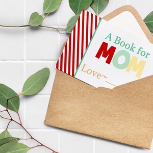 May include: A kraft paper envelope holds a card that reads "A Book for MOM" in red, green, and yellow. A red and white striped card is partially visible. Eucalyptus sprigs frame the scene, set against a white tiled background.