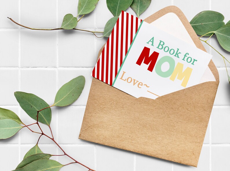 May include: A kraft paper envelope holds a card that reads "A Book for MOM" in red, green, and yellow. A red and white striped card is partially visible. Eucalyptus sprigs frame the scene, set against a white tiled background.