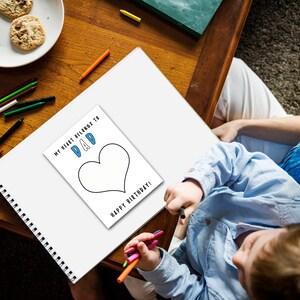 May include: A child colors a heart-shaped drawing on a white sheet of paper. The drawing says "My heart belongs to Dad" and "Happy Birthday!" The child is using colored pencils and is sitting at a table with a parent.