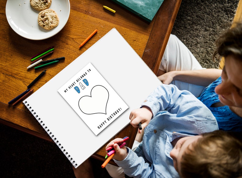 May include: A child colors a heart-shaped drawing on a white sheet of paper. The drawing says "My heart belongs to Dad" and "Happy Birthday!" The child is using colored pencils and is sitting at a table with a parent.