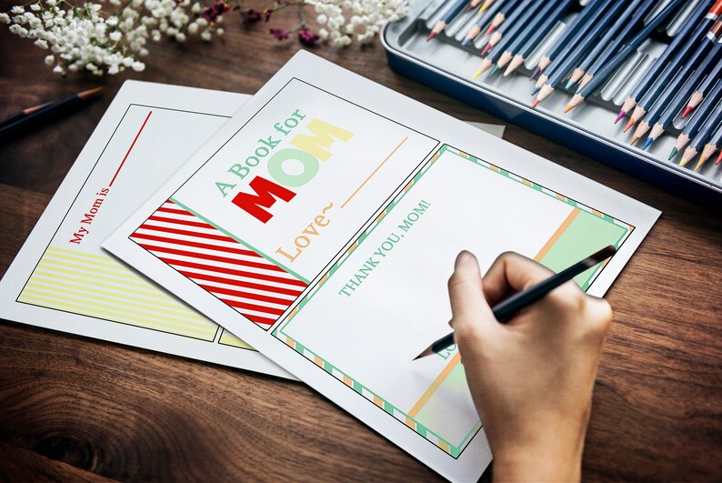 May include: A Book for Mom printable pages with colorful text and decorative borders. The pages include prompts like "My Mom is" and "Thank You, Mom!" A hand is writing with a pencil on one of the pages. Colored pencils are in a metal tin.