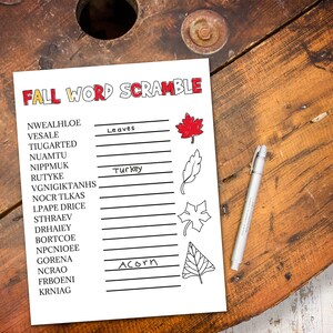 May include: A white sheet of paper with the words "FALL WORD SCRAMBLE" in red and orange. The paper has a list of scrambled words with blanks to unscramble. A pen rests on the right side of the paper. The background is a wooden surface.