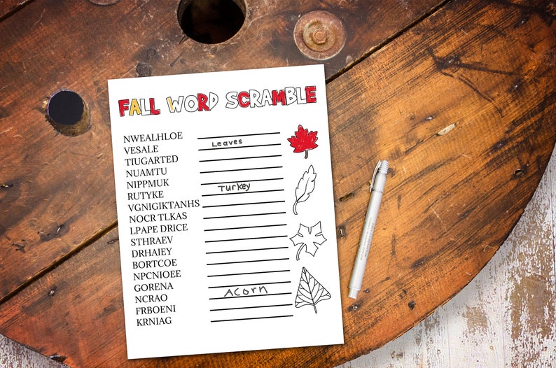 May include: A white sheet of paper with the words "FALL WORD SCRAMBLE" in red and orange. The paper has a list of scrambled words with blanks to unscramble. A pen rests on the right side of the paper. The background is a wooden surface.