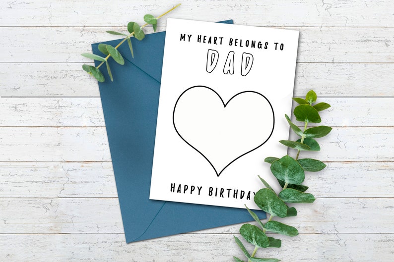 May include: A white card with black text that reads "My heart belongs to Dad" with a large heart outline below the text. The text "Happy Birthday" is below the heart. The card is on a white wooden surface with a blue envelope and green leaves.
