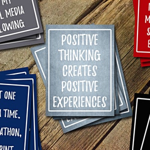 May include: A set of motivational cards with positive messages. The cards are blue, red, black, and gray with white text. The text on the blue card reads "Take it one step at a time. It's a marathon, not a sprint". The text on the red card reads "In, even means you're standing by yours". The text on the gray card reads "Positive thinking creates positive experiences".