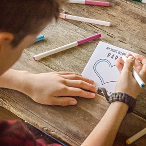 May include: A child colors a heart on a card that says "My heart belongs to Dad". The card is on a wooden table with colorful markers scattered around.