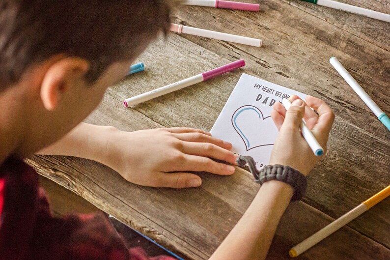 May include: A child colors a heart on a card that says "My heart belongs to Dad". The card is on a wooden table with colorful markers scattered around.