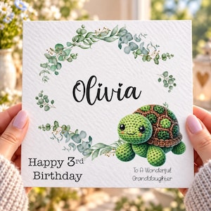 May include: A square birthday card featuring a green crocheted turtle, a watercolour eucalyptus wreath, and the text Olivia, Happy 3rd Birthday, and To A Wonderful Granddaughter printed in black on a white textured background.