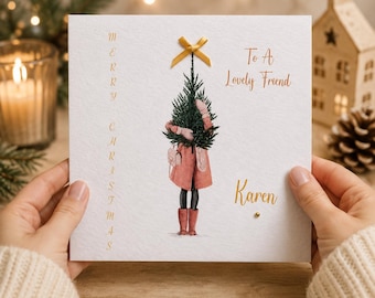 Könnte beinhalten: Eine quadratische weiße Grußkarte mit goldener Schrift, die links vertikal Merry Christmas und rechts To A Lovely Friend Karen lautet. Eine zentrale Illustration zeigt eine Figur in einem rosa Mantel, die einen grünen Tannenbaum hält.
