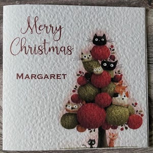 May include: A Christmas card featuring the text "Merry Christmas" and the name "Margaret". The card's design includes a Christmas tree made of red and green yarn balls, with cats incorporated into the design. The background is textured and off-white.