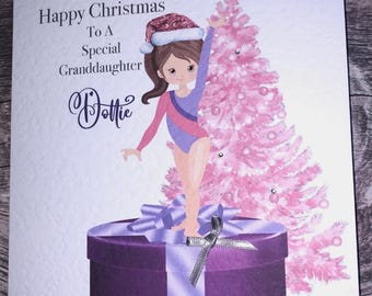 Può includere: Un biglietto di Natale con una ginnasta in piedi su una scatola regalo viola con un albero di Natale rosa dietro di lei. Il biglietto dice "Buon Natale a una nipote speciale Dottie".
