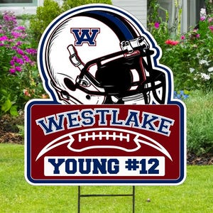May include: A yard sign featuring a football helmet and the text "WESTLAKE YOUNG #12". The helmet is white with a blue and black design. The sign is red and blue, and is placed in a grassy yard.
