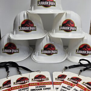 May include: Six white hard hats with the Jurassic Park logo printed on them. Five of the hats have the logo in black and red, and one hat has the logo in black and green. There are five white plastic ID badges with black lanyards. The badges have the Jurassic Park logo and the text "Park Ranger" and "Licensed Dinosaur Expert".