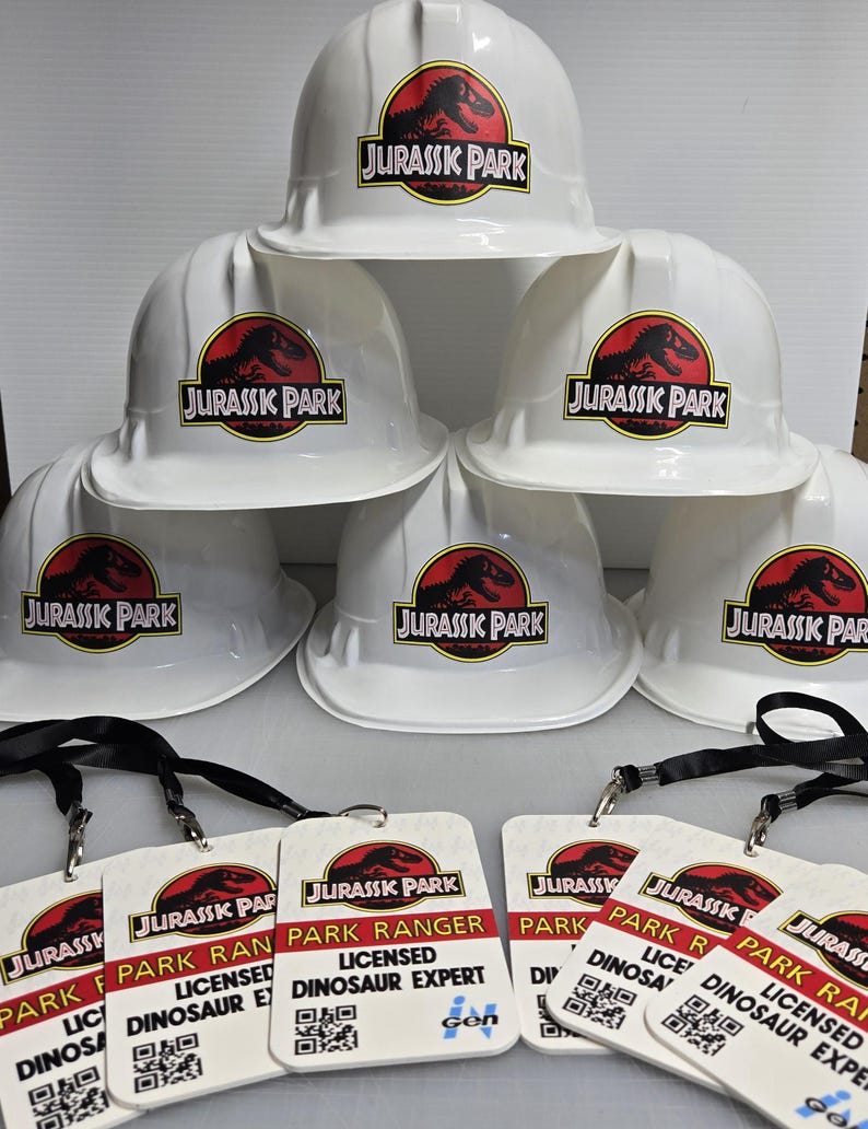 May include: Six white hard hats with the Jurassic Park logo printed on them. Five of the hats have the logo in black and red, and one hat has the logo in black and green. There are five white plastic ID badges with black lanyards. The badges have the Jurassic Park logo and the text "Park Ranger" and "Licensed Dinosaur Expert".