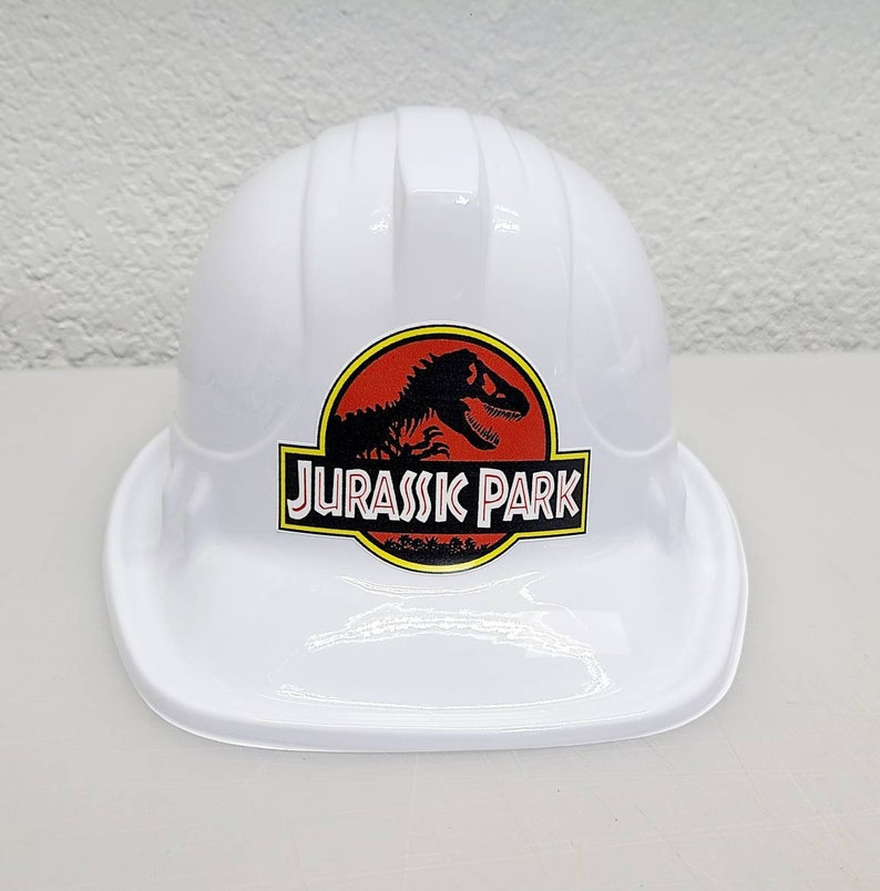 May include: White plastic construction worker hard hat with a red and yellow Jurassic Park logo featuring a black silhouette of a dinosaur.