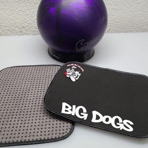May include: A purple bowling ball with a black base and the word "HAMMER" printed on it. Two bowling accessories are in the foreground: a black mat with the words "BIG DOGS" and a cartoon dog, and a gray mat with black dots.