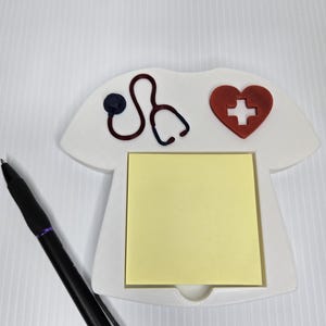 May include: A white, nurse-themed memo pad holder with a red heart and cross, and a stethoscope design. A black pen with purple accents rests beside the holder, which contains a yellow sticky note pad.