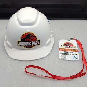 May include: White hard hat with a Jurassic Park logo featuring a dinosaur and the text "Jurassic Park". A white ID card with the text "Jurassic Park Park Ranger Licensed Dinosaur Expert" and a red lanyard.