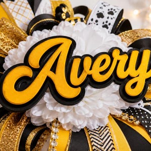 May include: A decorative rosette featuring the name Avery in yellow script lettering with a black outline, centered on a white chrysanthemum flower. The base is layered with black, yellow, and white ribbons, including patterns of paw prints and gold glitter.