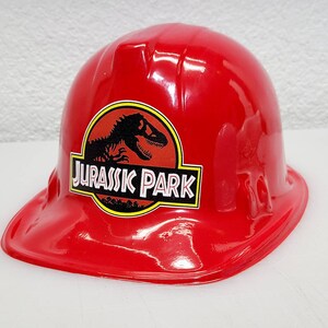 May include: Red plastic construction worker hat with a Jurassic Park logo featuring a dinosaur silhouette. The logo is black and yellow with the words "Jurassic Park" in white.