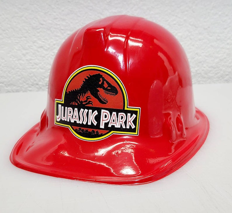 May include: Red plastic construction worker hat with a Jurassic Park logo featuring a dinosaur silhouette. The logo is black and yellow with the words "Jurassic Park" in white.