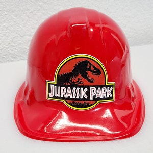 May include: Red plastic construction worker hat with a Jurassic Park logo featuring a dinosaur silhouette and the text "Jurassic Park".