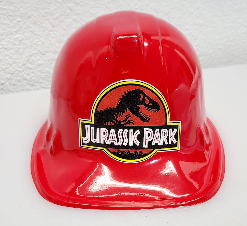 May include: Red plastic construction worker hat with a Jurassic Park logo featuring a dinosaur silhouette and the text "Jurassic Park".