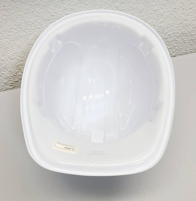 May include: White hard hat with a smooth, glossy finish. The inside of the hard hat is visible, showing the suspension system. The hard hat has a label that reads "WARNING".