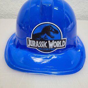May include: A blue plastic construction worker hat with a Jurassic World logo featuring a dinosaur silhouette.
