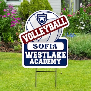 May include: A volleyball yard sign with a volleyball graphic and the word "VOLLEYBALL" in a red banner. The sign also includes the text "SOFIA WESTLAKE ACADEMY" in white letters on a blue background. The sign is displayed on a green lawn.
