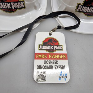 May include: A white plastic hard hat with the words "Jurassic Park" printed in red and black. A black lanyard with a white plastic badge attached. The badge has a red and black Jurassic Park logo, the words "Park Ranger", "Licensed Dinosaur Expert", and a QR code. The badge also has the words "iGen" printed in blue and white.