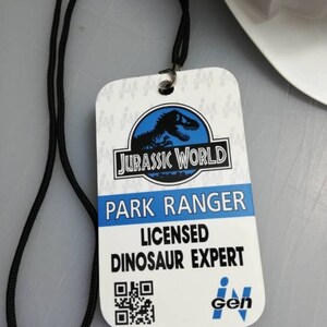 May include: A white plastic ID card with a black lanyard. The card has a blue Jurassic World logo with a dinosaur silhouette. The text on the card reads "Park Ranger Licensed Dinosaur Expert" and has a black and white QR code. The card also has a blue InGen logo.