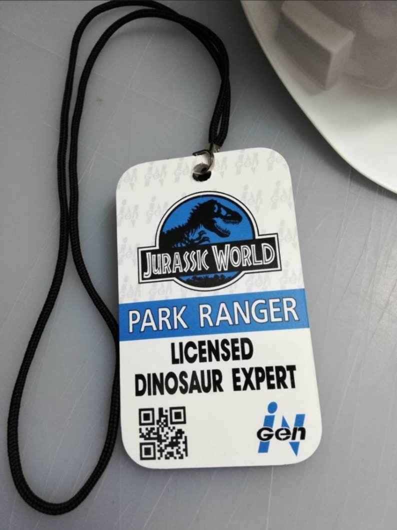 May include: A white plastic ID card with a black lanyard. The card has a blue Jurassic World logo with a dinosaur silhouette. The text on the card reads "Park Ranger Licensed Dinosaur Expert" and has a black and white QR code. The card also has a blue InGen logo.