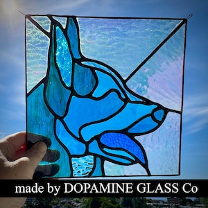 May include: A square stained glass panel featuring a blue dog silhouette with pointed ears and an open mouth. The design uses various shades of blue glass with black lead lines. The text "made by DOPAMINE GLASS Co" is printed at the bottom.
