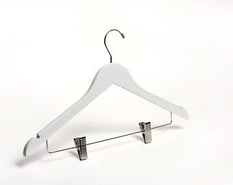 May include: White wooden hanger with a chrome hook and two metal clips. The hanger is designed for hanging clothes with a built-in bar for holding pants or skirts.