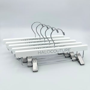May include: Set of five white wooden hangers with silver metal clips and the text "HALOCOULTURE" printed on the front of each hanger.