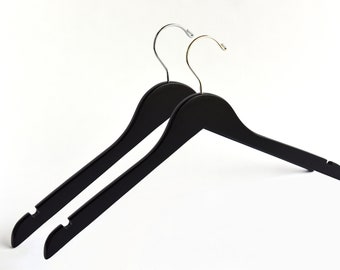 May include: Two black wooden clothes hangers with silver and gold-colored hooks. The hangers are designed with a curved shoulder shape and a notched bar for securing garments. The background is plain white.