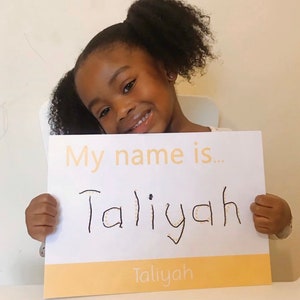 May include: A young girl holds up a white sign with the words "My name is..." and "Taliyah" written in black marker. The name "Taliyah" is written in a yellow banner at the bottom of the sign.