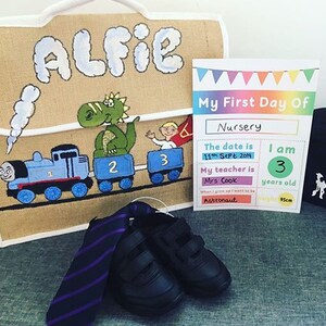 May include: A brown burlap bag with the name "Alfie" painted on it. The bag is decorated with a green dinosaur, a blue train, and the numbers 1, 2, and 3. A printed card with the text "My First Day Of Nursery" is next to the bag. The card includes the date, the child's age, and the teacher's name. A black tie and two black shoes are in the foreground.
