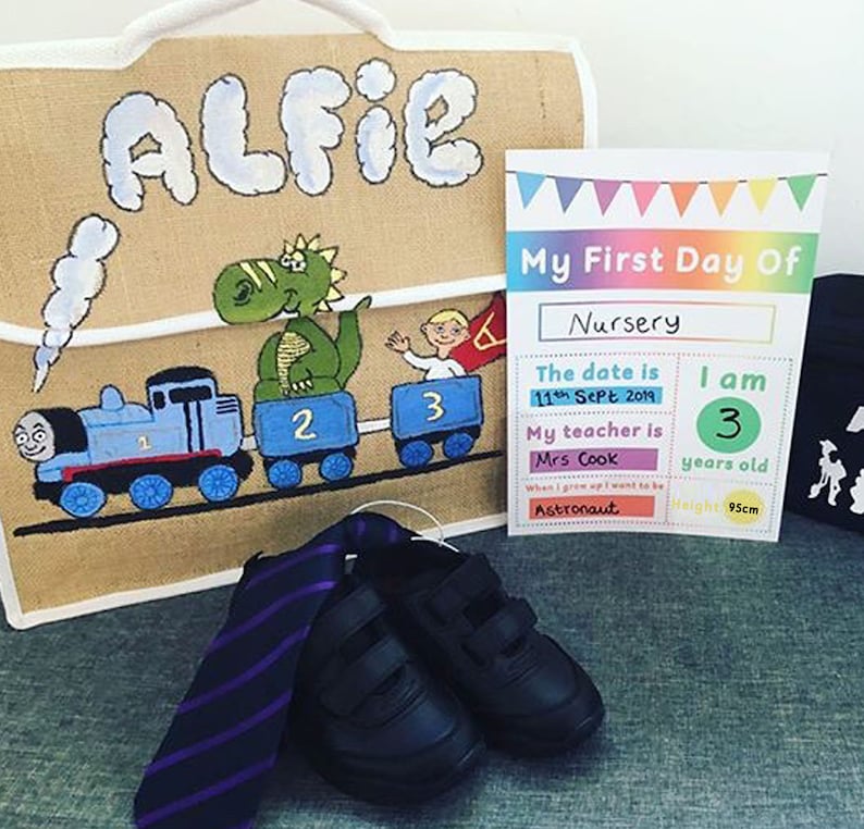 May include: A brown burlap bag with the name "Alfie" painted on it. The bag is decorated with a green dinosaur, a blue train, and the numbers 1, 2, and 3. A printed card with the text "My First Day Of Nursery" is next to the bag. The card includes the date, the child's age, and the teacher's name. A black tie and two black shoes are in the foreground.