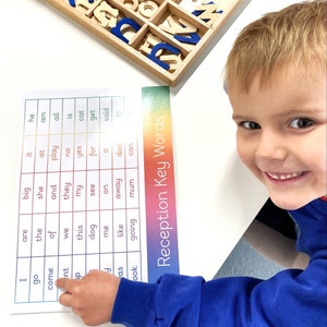 May include: A young child points to a word on a colorful chart with the title "Reception Key Words". The chart lists common words used in early literacy education. The child is wearing a blue shirt.