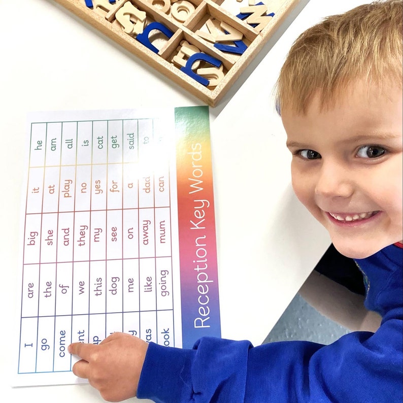 May include: A young child points to a word on a colorful chart with the title "Reception Key Words". The chart lists common words used in early literacy education. The child is wearing a blue shirt.