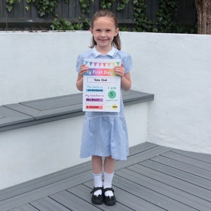 May include: A young girl in a blue and white plaid dress holds a sign that says "My First Day" with the date "05/04/2019" and the text "I am 5 years old".