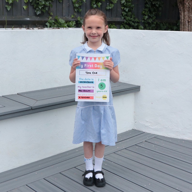 May include: A young girl in a blue and white plaid dress holds a sign that says "My First Day" with the date "05/04/2019" and the text "I am 5 years old".