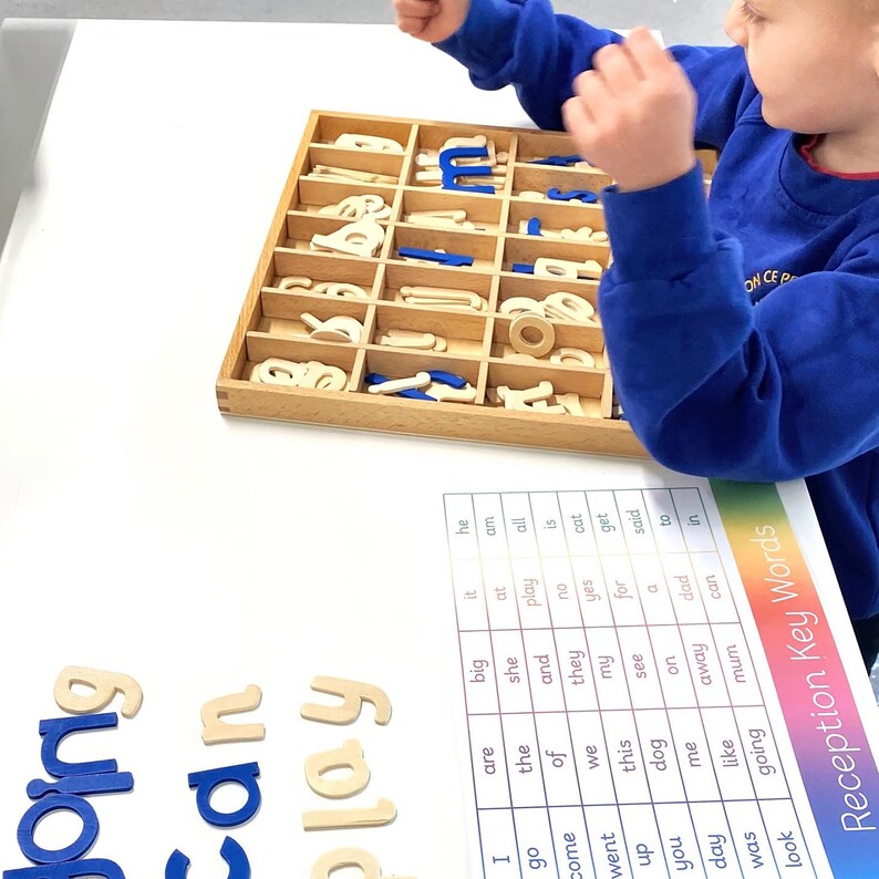 May include: A child is learning to spell using a wooden letter tray and a chart of common words. The tray contains a variety of wooden letters in both uppercase and lowercase. The chart lists words such as "big", "play", "cat", "see", "dog", "mum", and "look".