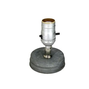 Pode incluir: Uma pequena base de lâmpada cinza com um soquete de lâmpada prateado. A base é redonda e feita de um material texturizado. O soquete tem um interruptor liga/desliga preto e um interior cor de latão.