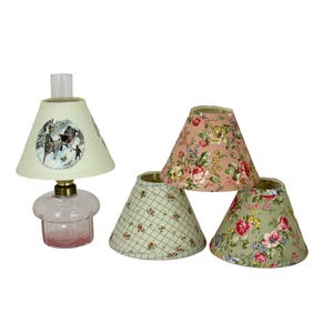 May include: A small, pink glass lamp with a cream-colored lampshade featuring a winter scene. Three additional lampshades are included, with floral and plaid patterns in pink, green, and cream colors. The lamp has a brass base.