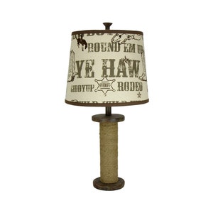 May include: A rustic table lamp with a brown rope base and a beige lampshade with a western-themed design. The lampshade features the words "Ye Haw", "Round Em Up", "Rodeo", and "Giddy Up" in brown text.