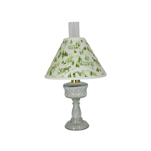May include: A white lampshade with a green and white country scene print sits atop a clear glass lamp base with a textured design.