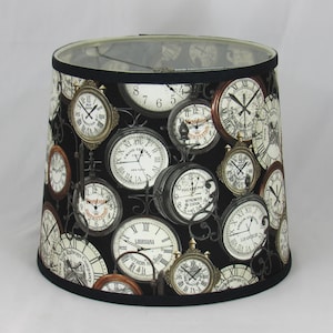 Pode incluir: Abajur cilíndrico com fundo preto adornado com vários desenhos de relógios antigos. Os relógios têm mostradores brancos com algarismos romanos e molduras de metal ornamentadas. As bordas superior e inferior são aparadas com tecido preto.